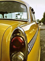 Close-up of a yellow vintage car’s rear side and taillight with a black-and-white checkered stripe.