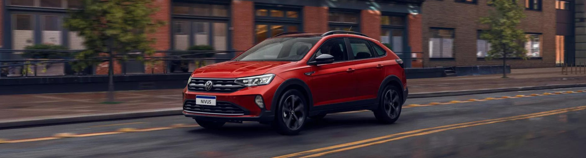 SUV vermelho trafegando em uma rua da cidade, com prédios de tijolos e árvores ao fundo.