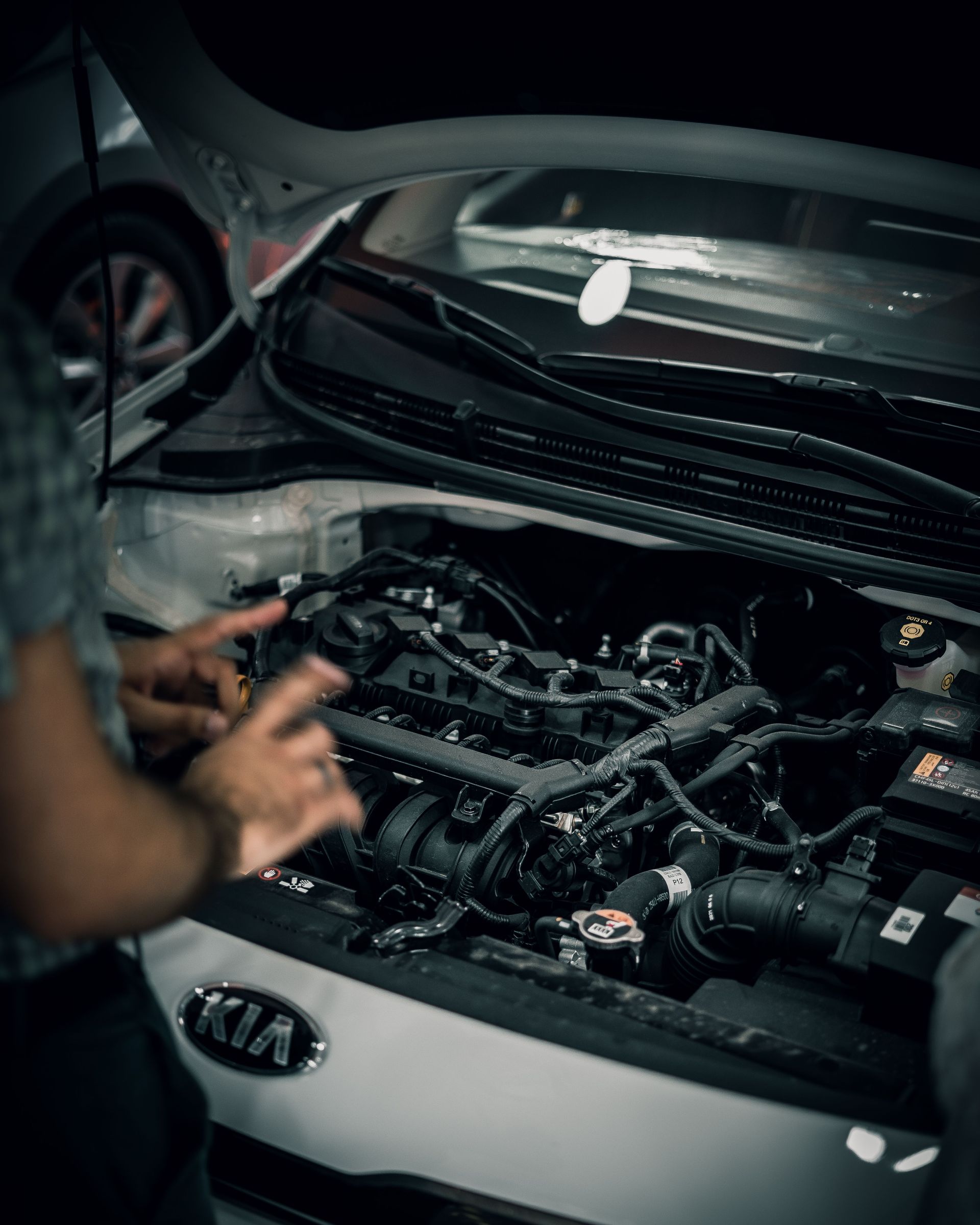Mecánico trabajando en el motor de un coche Kia con el capó abierto en un garaje con poca luz.