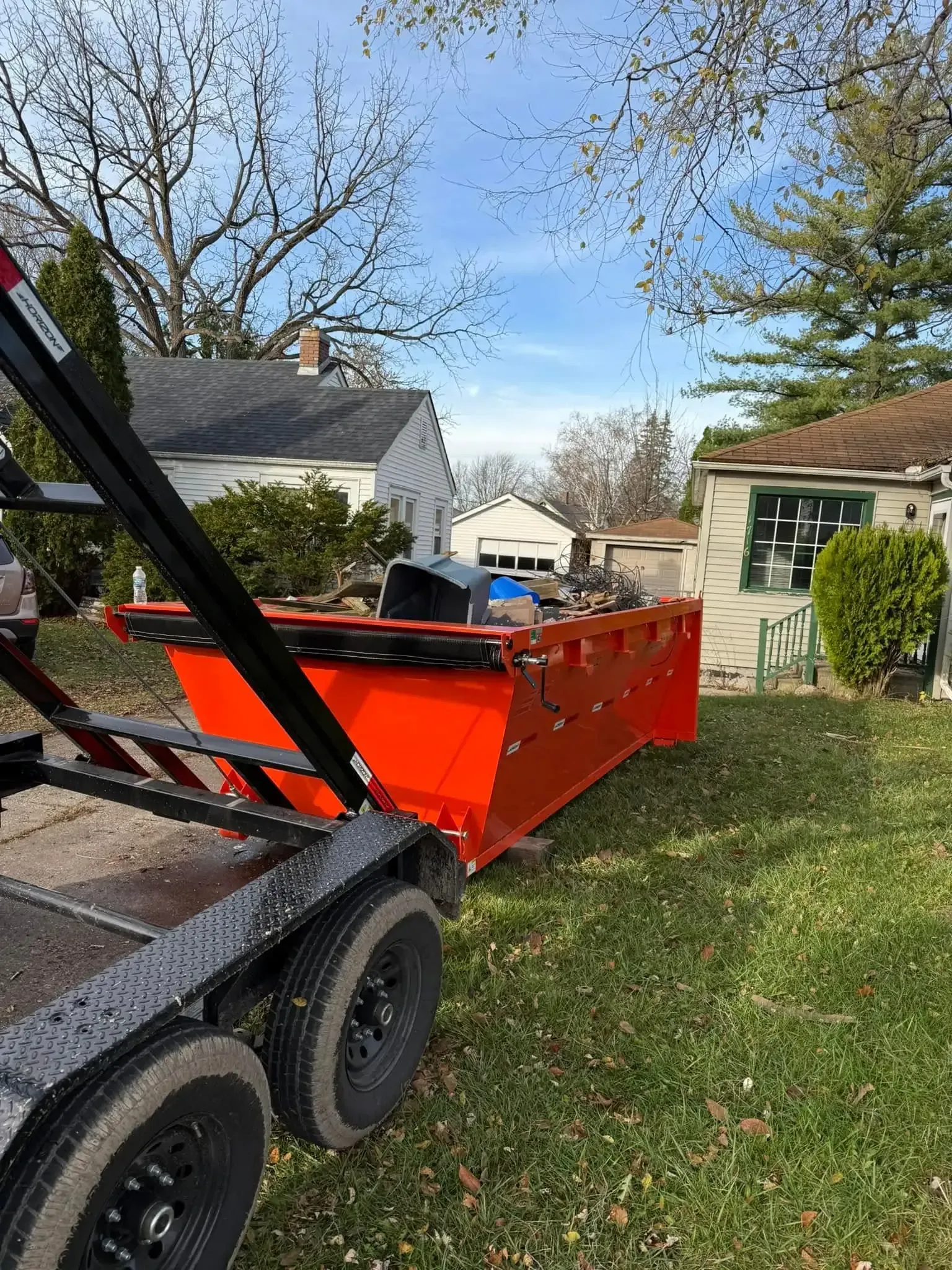 Orange dump trailer in a yard beside a house under a clear blue sky