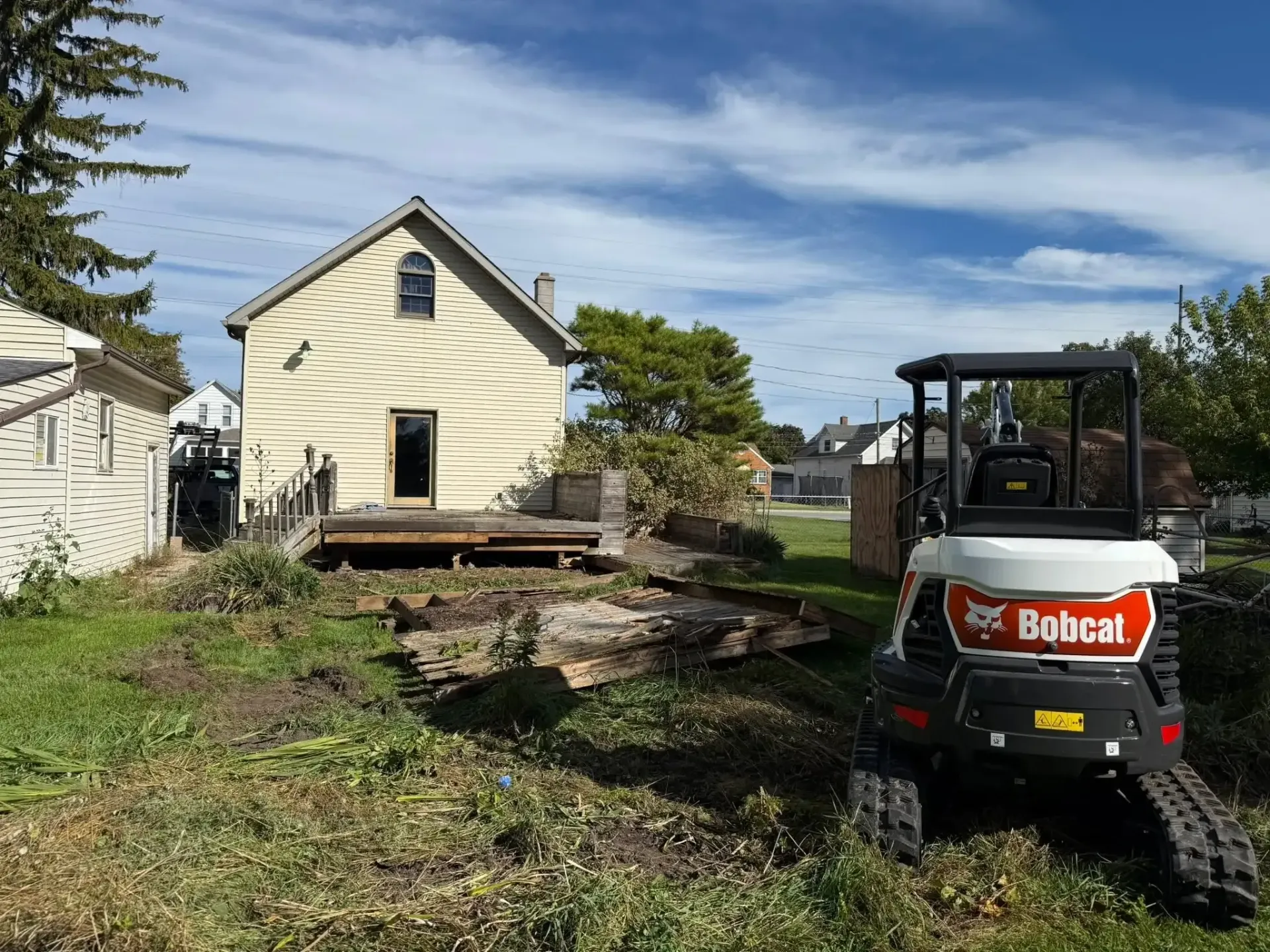 Bobcat skid steer in yard beside a cream house under construction, with dirt pile and cleared ground.