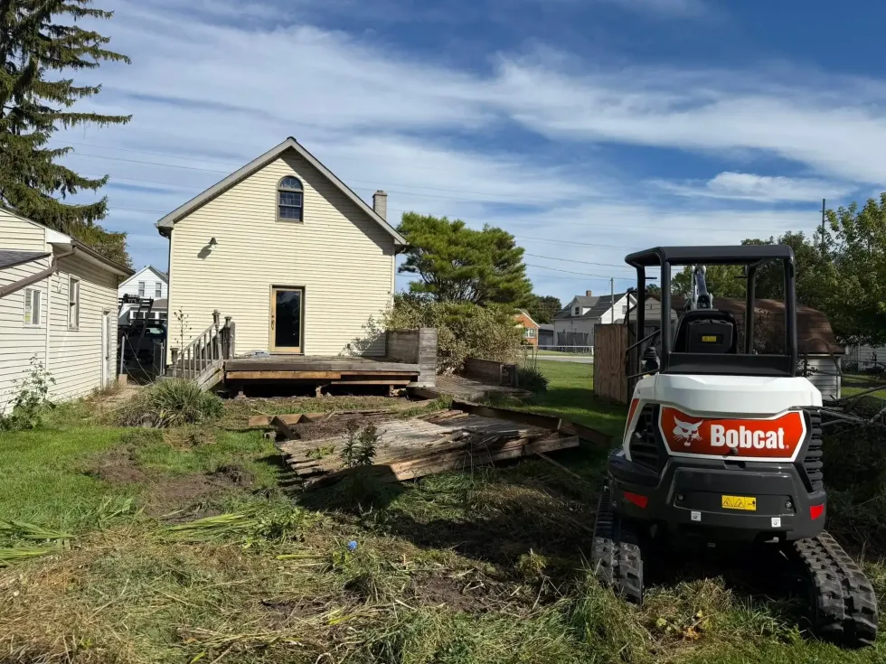 Bobcat skid steer in yard beside a cream house under construction, with dirt pile and cleared ground.