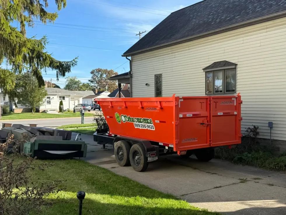 Orange dumpster trailer parked beside a cream house on a driveway, with yard and street in the background.