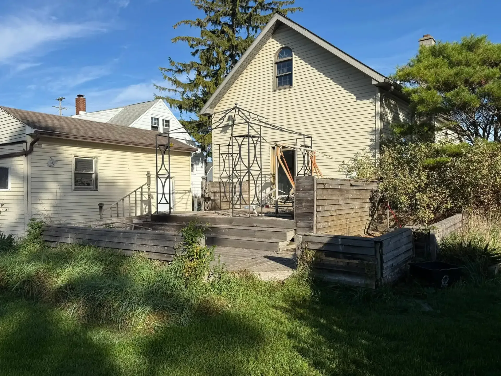 Backyard patio with stone steps, small metal arbor, beige house, and a large tree in sunlight