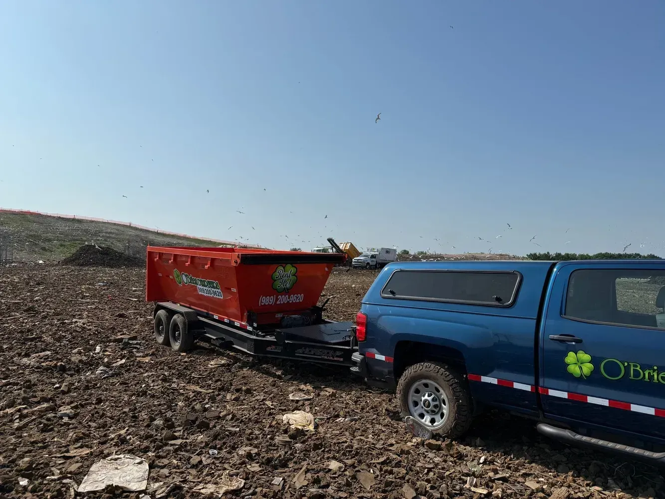 Blue pickup towing a red dumpster trailer across a rocky landfill under a clear sky