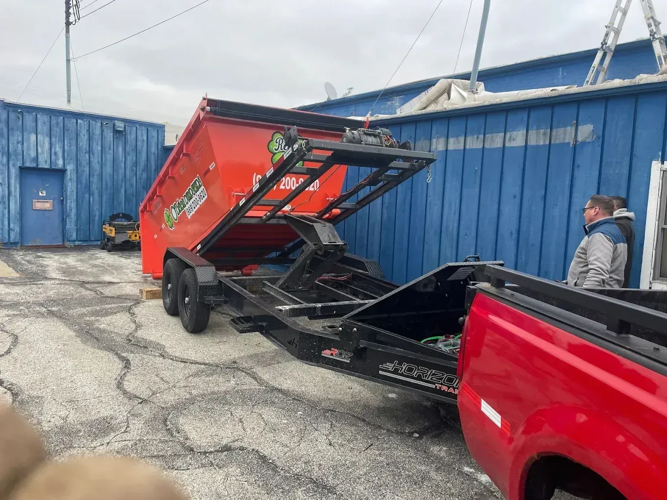 Dumpster being lifted by a red truck beside blue shipping containers in an industrial yard