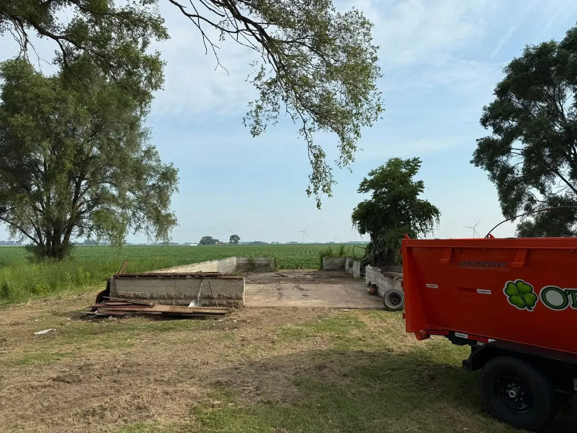 Rural field with trees, a dirt area, and a red trailer at the right edge under a cloudy sky