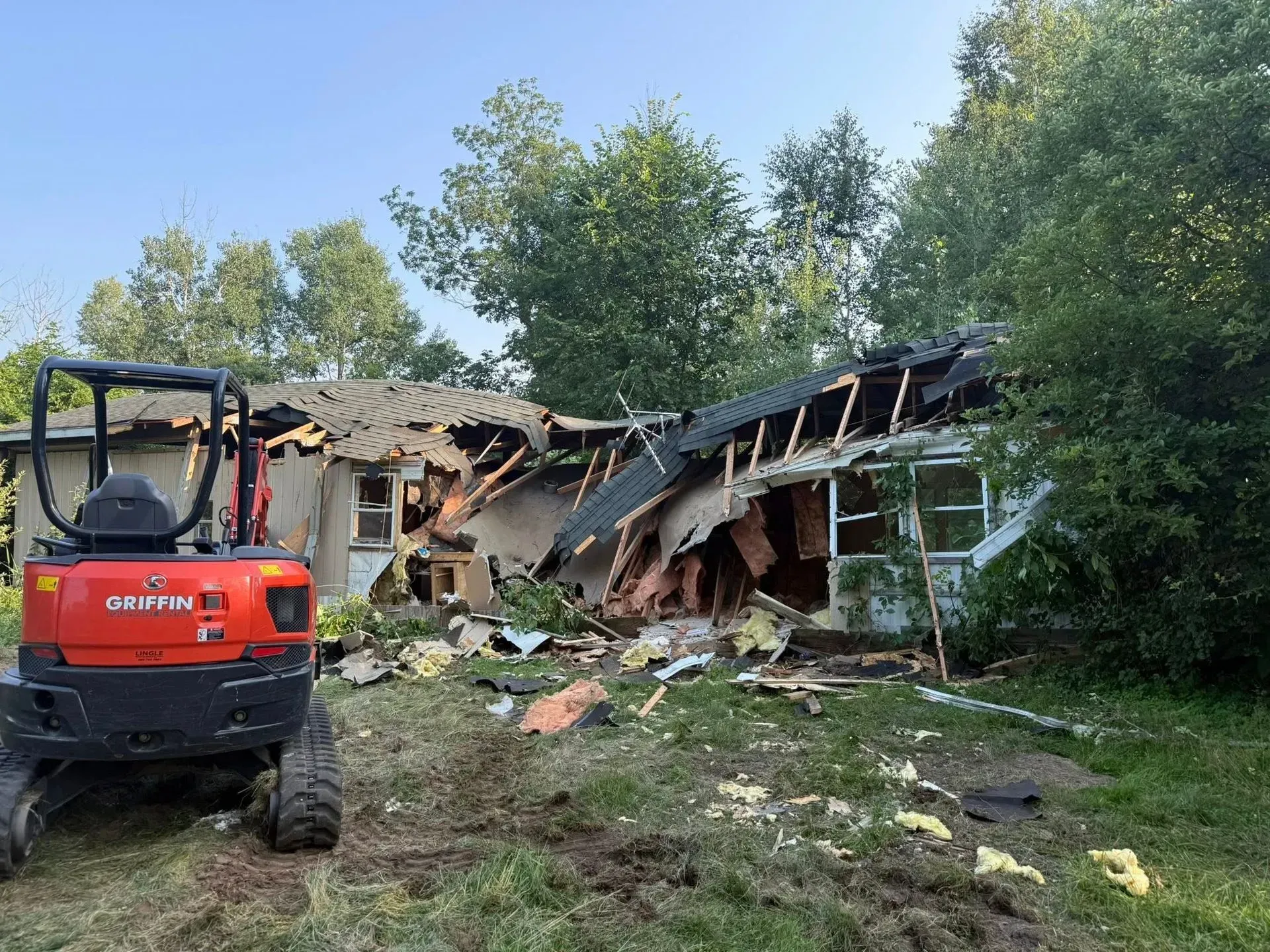 Red excavator beside a partially collapsed house with debris and trees in the yard