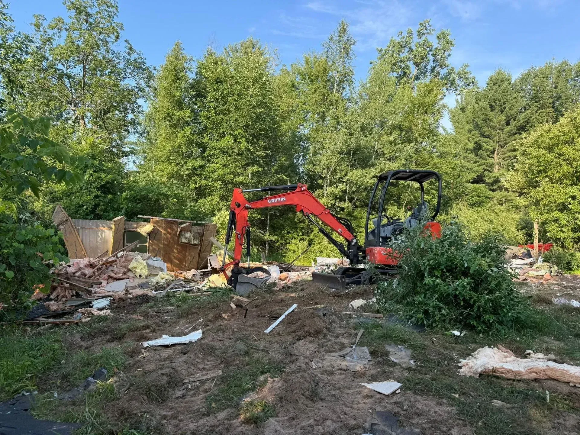 Red excavator clearing debris in a wooded area under a blue sky.