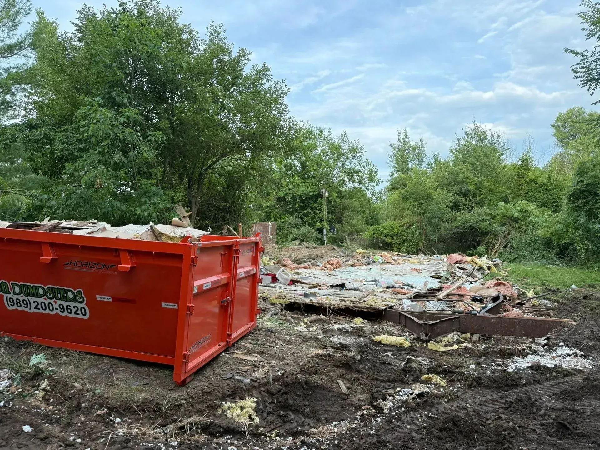 Red dumpster beside a muddy demolition site with rubble and green trees under a cloudy sky