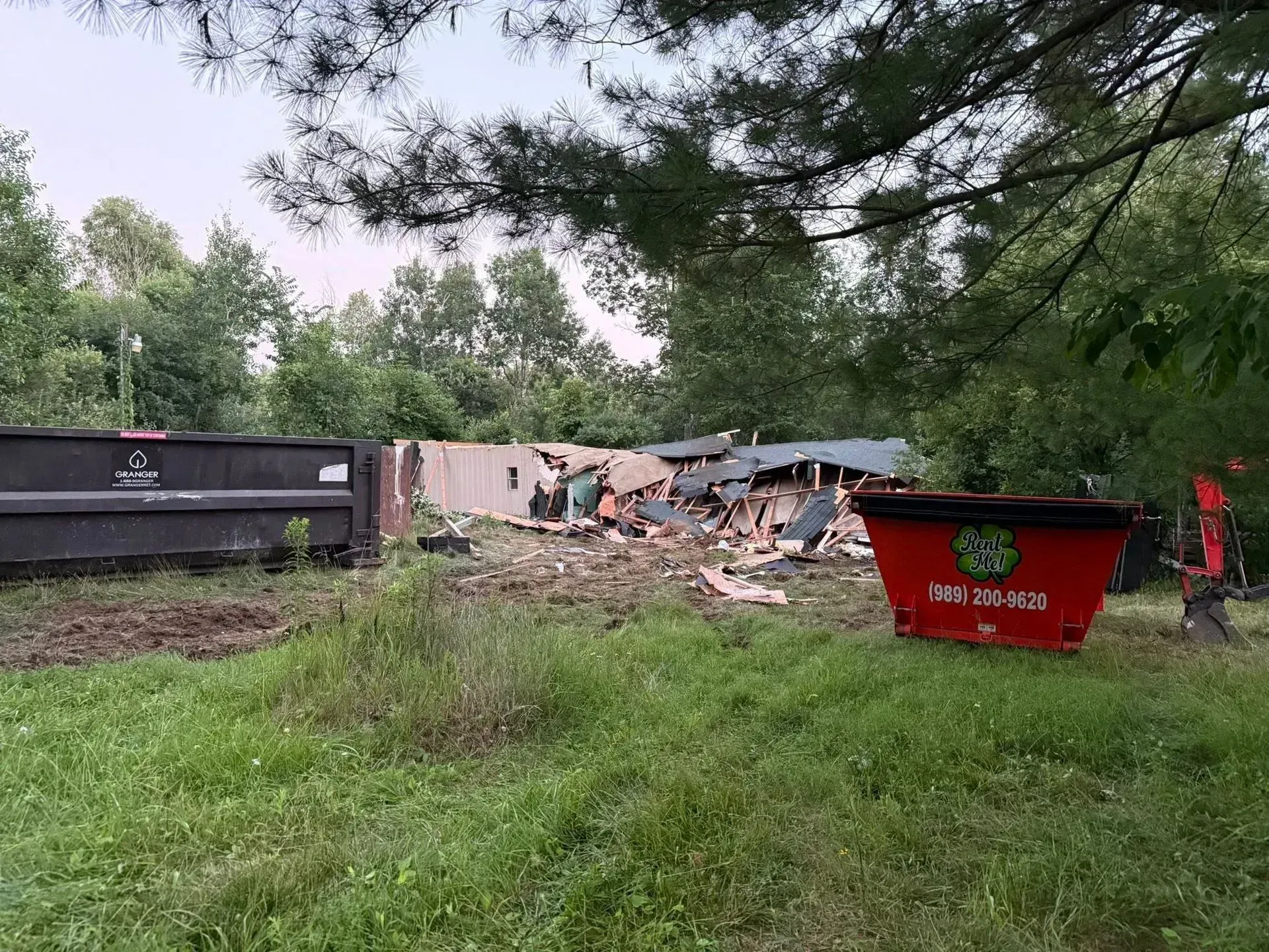 Demolition debris beside a red dumpster in a grassy yard with trees overhead