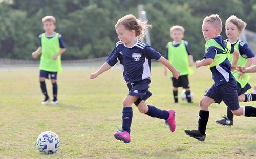Children playing soccer on a grassy field, with one dribbling a ball in the foreground.