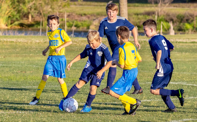Children playing soccer on a grassy field, with one in blue dribbling the ball amid players in yellow and blue jerseys.