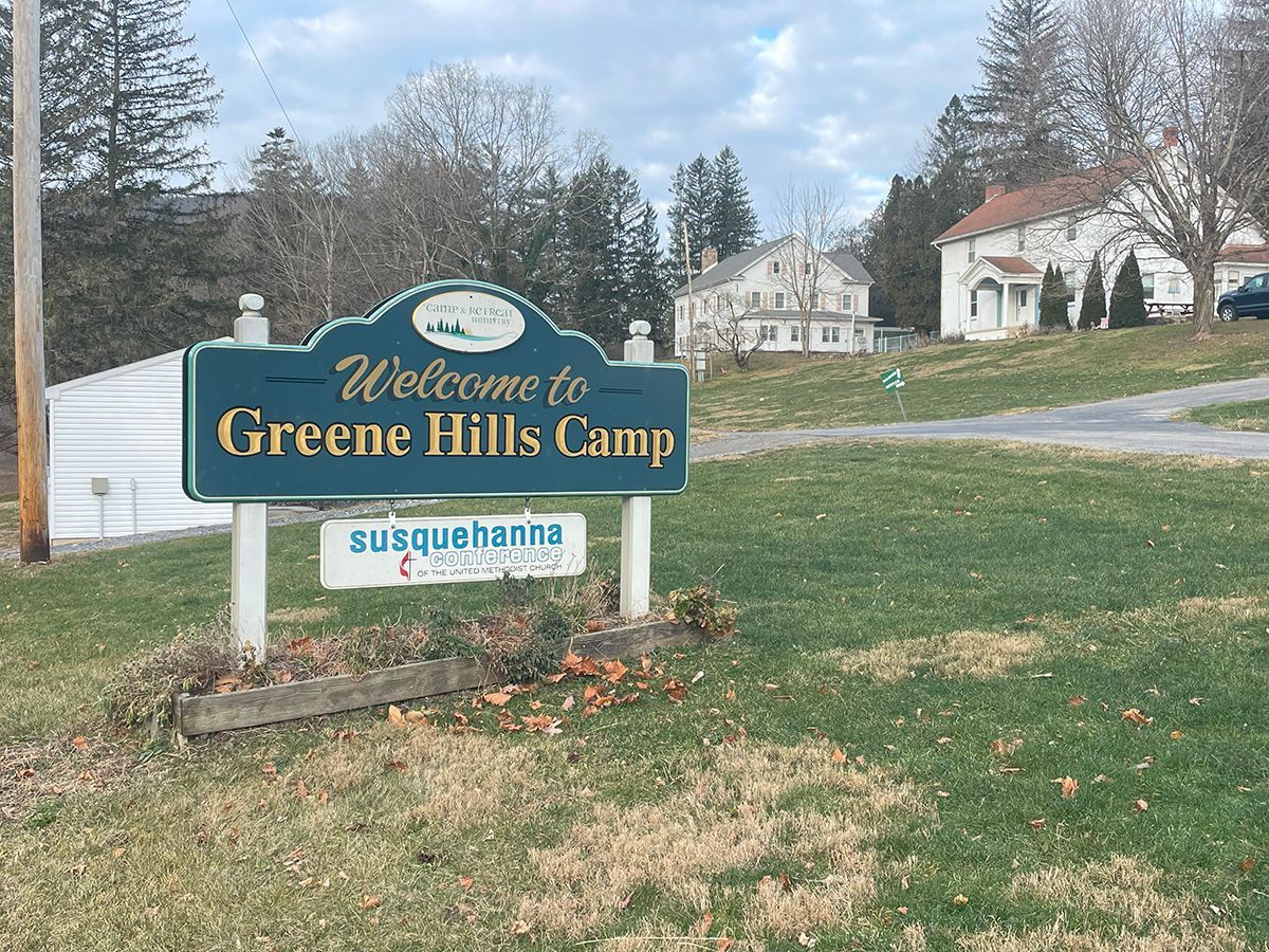 A welcome to greene hills camp sign is in the middle of a grassy field.