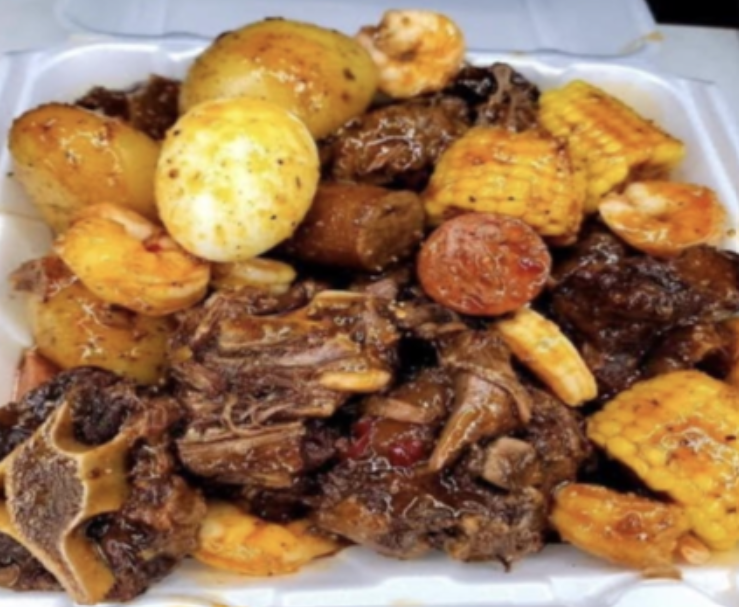 Foil tray of roasted meat, potatoes, corn, and sausage in dark seasoning sauce