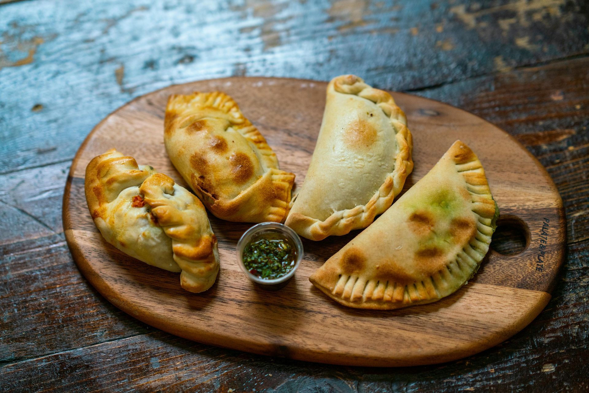 Assorted baked empanadas on a wooden board with a small bowl of green sauce