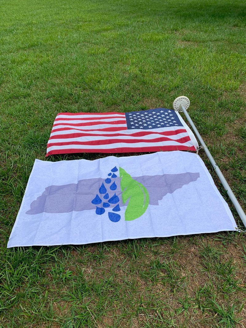 Two flags laid on grass: a U.S. flag above a white flag with a blue-green raindrop logo.