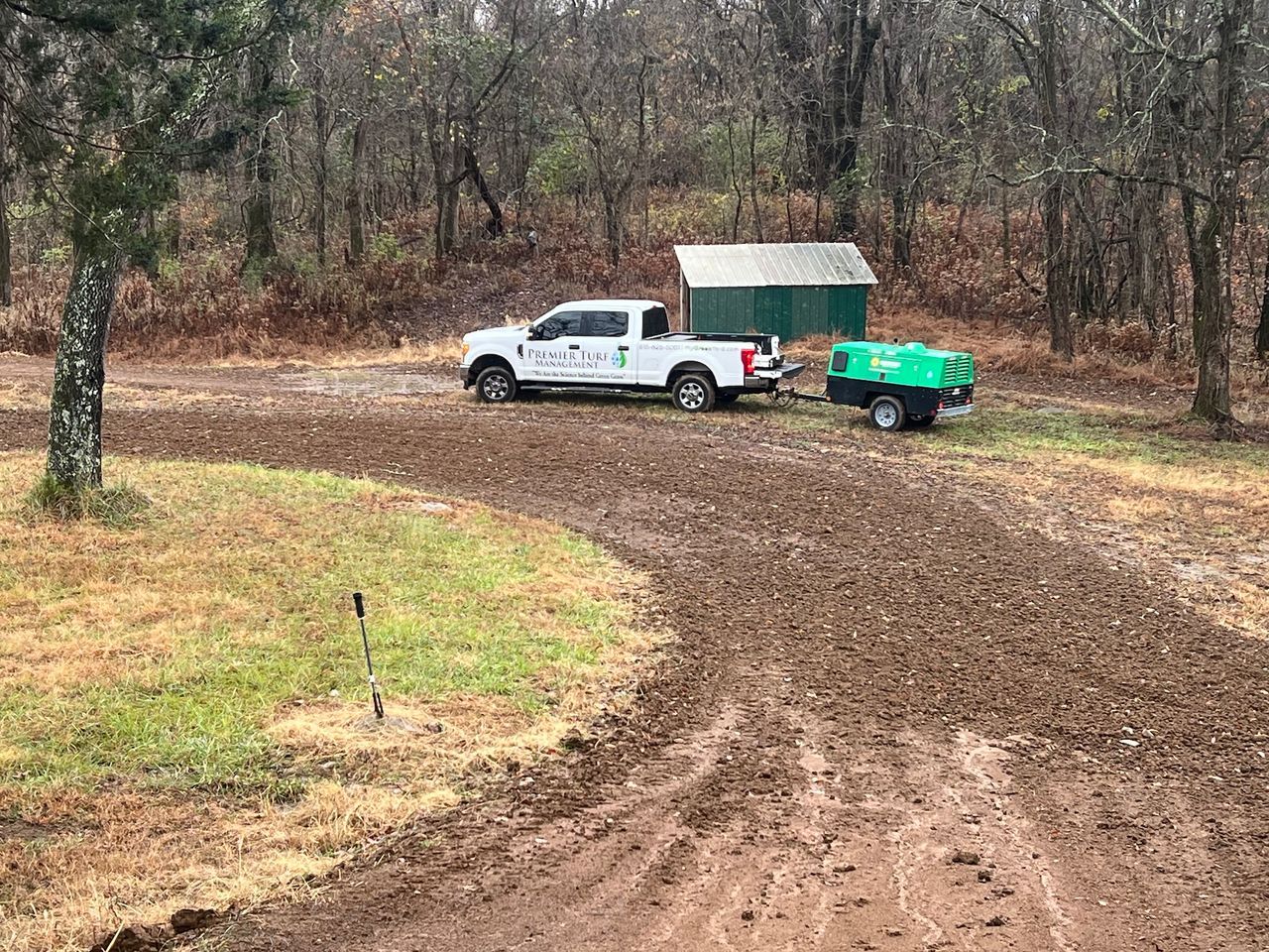 White pickup truck towing green trailer on a muddy dirt road in a wooded area.