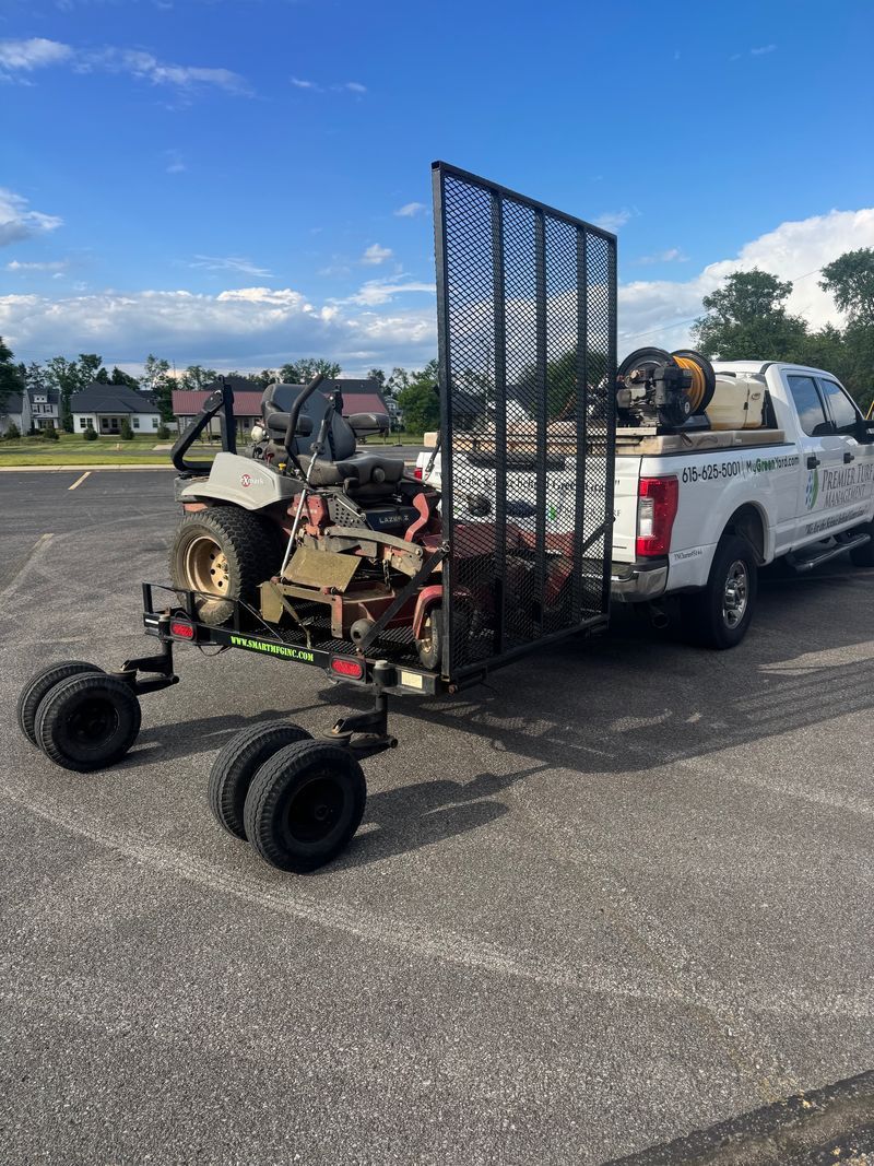 White pickup with a small trailer carrying a metal mesh ramp and a riding mower in a parking lot
