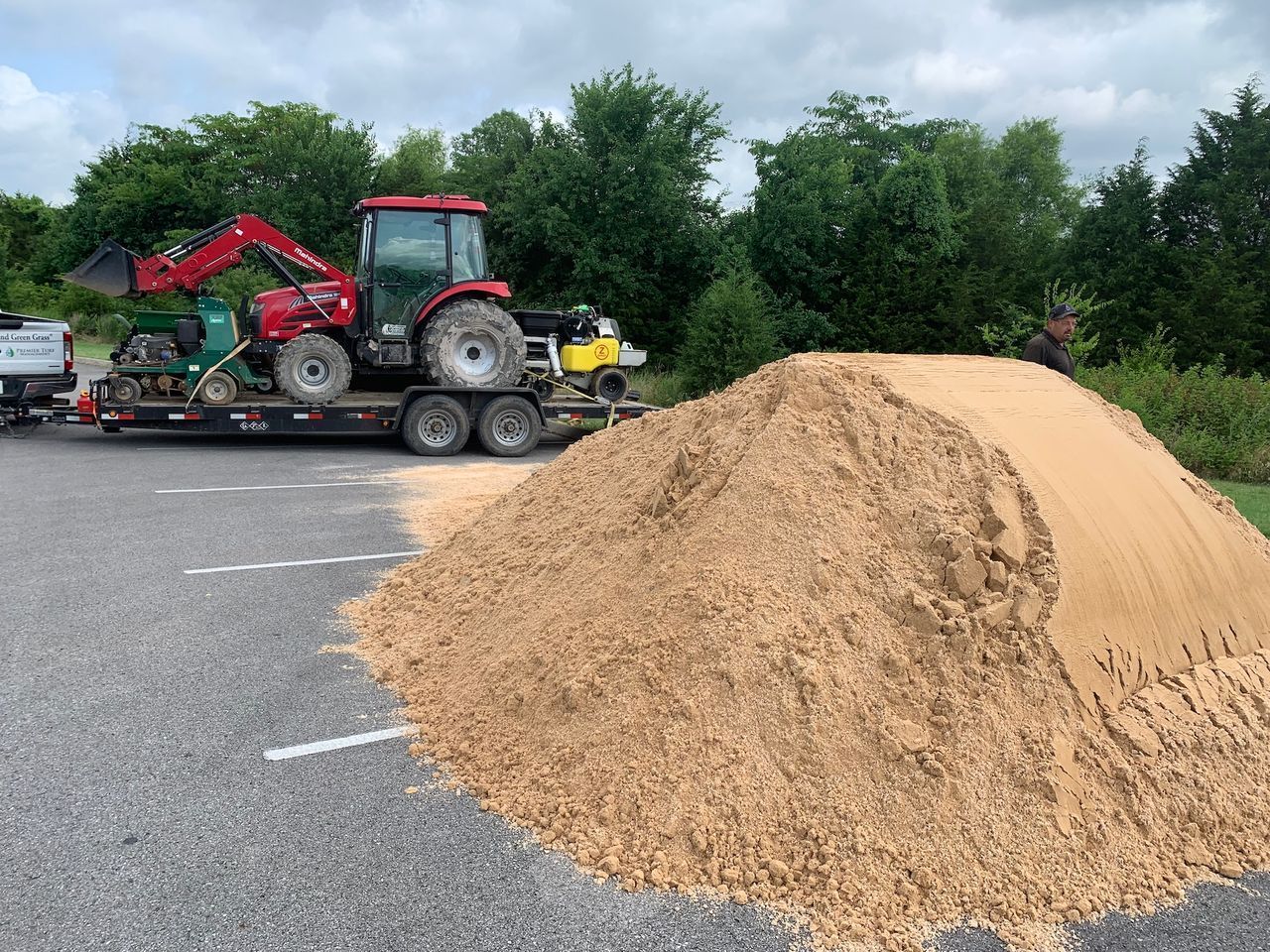 Red tractor hauling farm equipment beside a large pile of wood chips in a parking lot