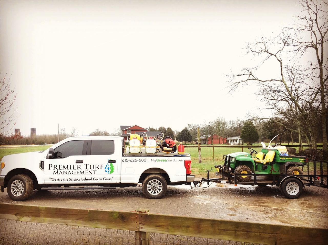 White pickup truck towing a green trailer on a wet road beside a grassy field and trees