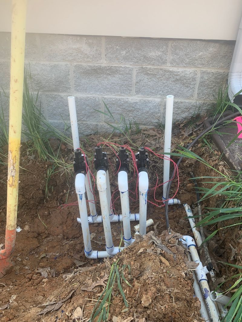 Outdoor utility pipes and valves beside a wall, partly buried in dirt and grass