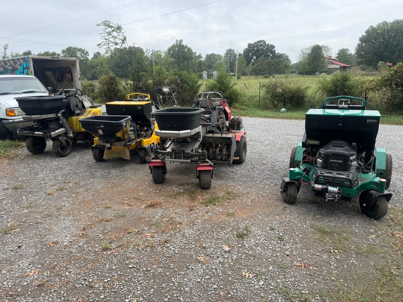 Three lawn tractors and a pickup truck parked on gravel in a rural yard