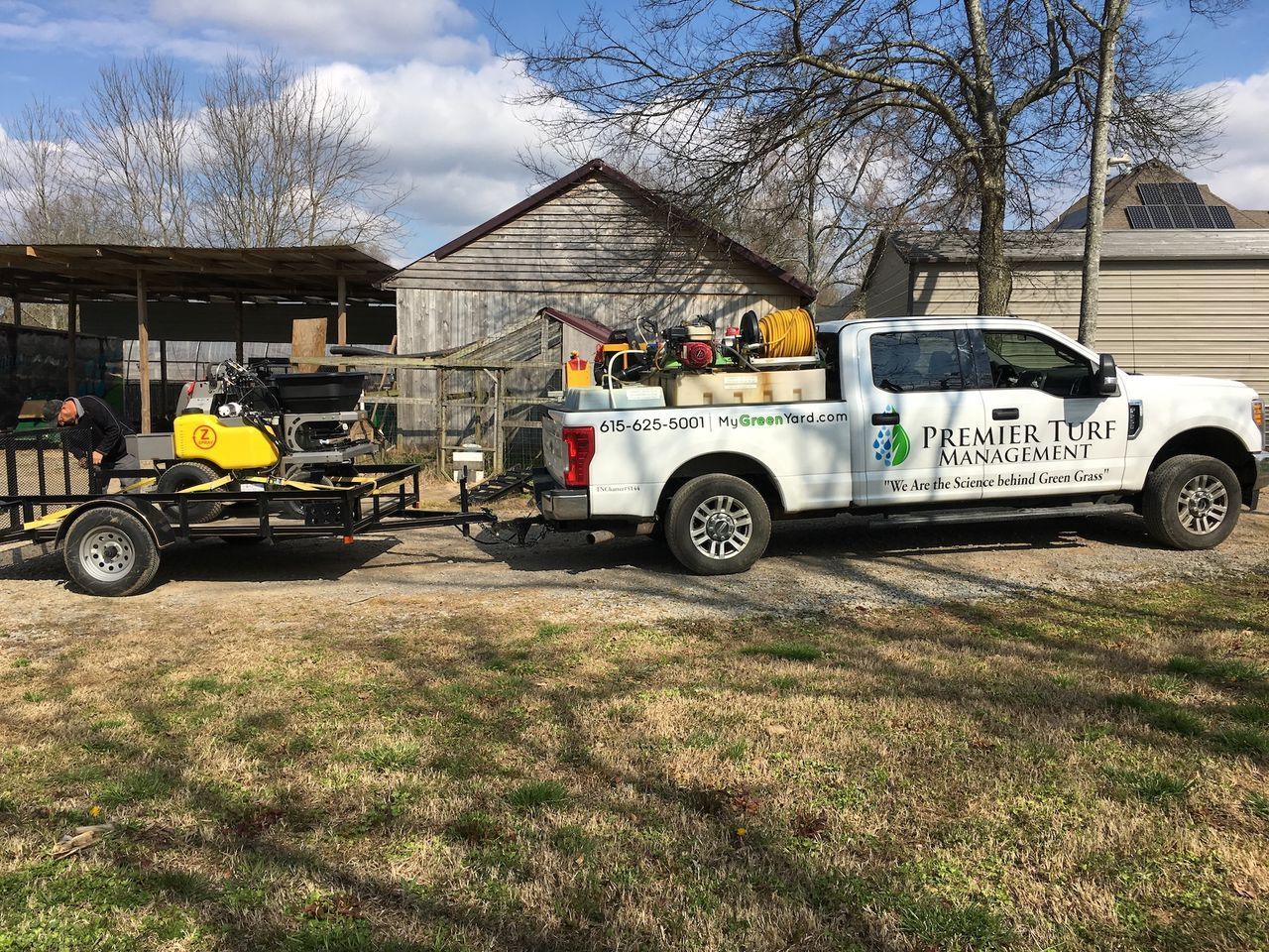 White utility truck towing a yellow excavator in a yard beside a house and shed