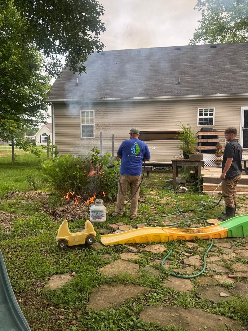 Two people use a leaf blower and hose near a house with smoke or mist rising from the roof.