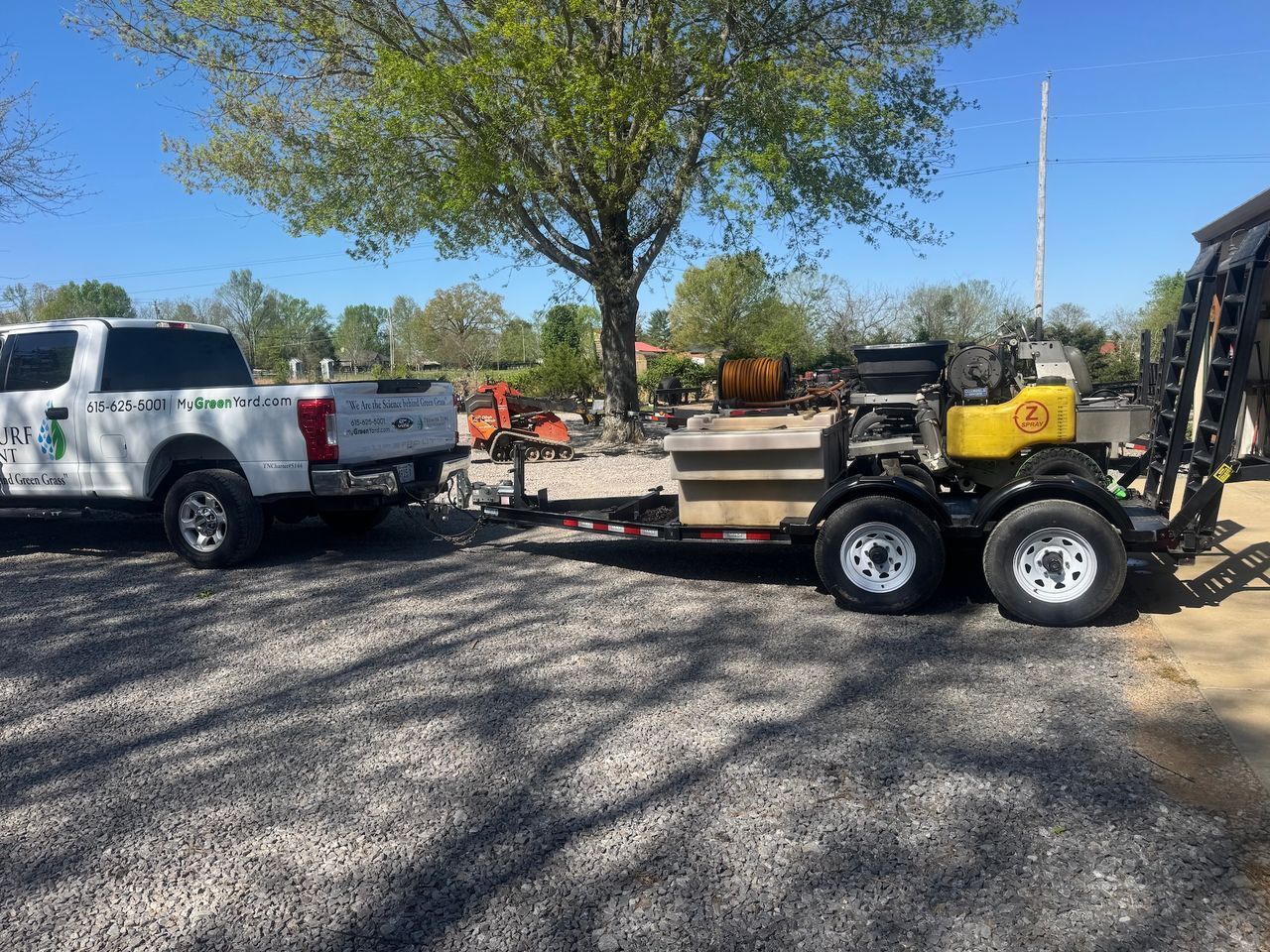 White pickup towing a yellow skid steer on a flatbed trailer in a gravel lot under a tree.