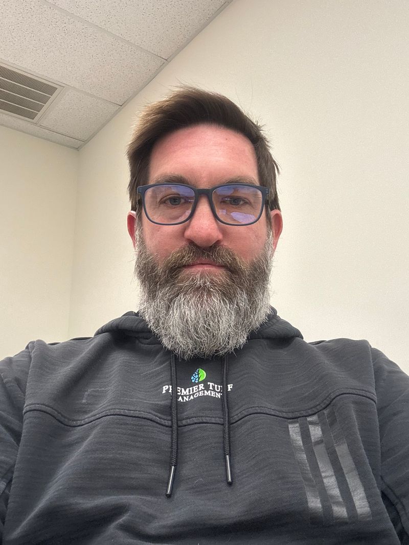 Man with glasses and gray beard wearing a dark hoodie, seated indoors under office lighting.