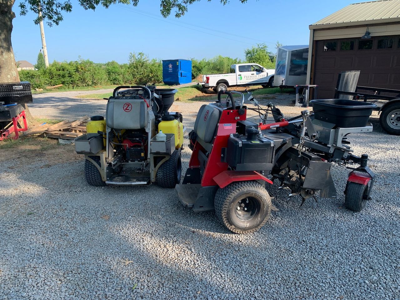 Two red riding lawn mowers parked on gravel near a building and truck outdoors.