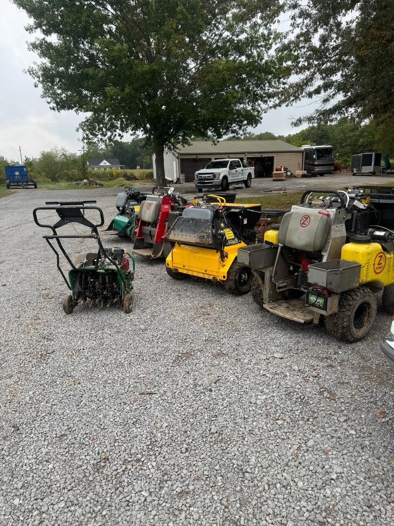 Outdoor yard with several small utility vehicles and equipment parked on gravel under a large tree
