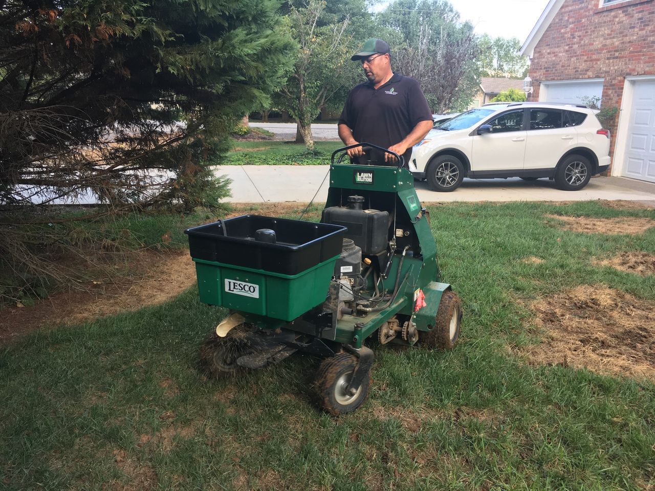 Person operating a green lawn machine on grass near a white SUV in a suburban yard