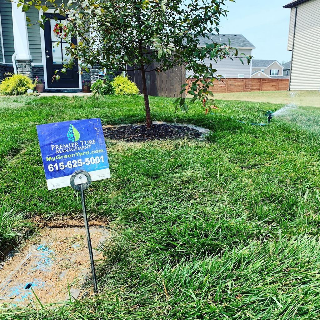 Blue lawn care sign in a green yard beside a small tree and suburban house.