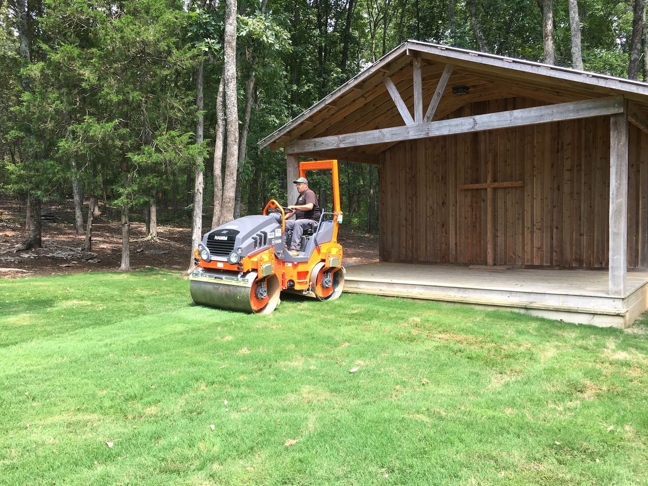 Orange riding mower on grass beside a wooden shed in a wooded yard