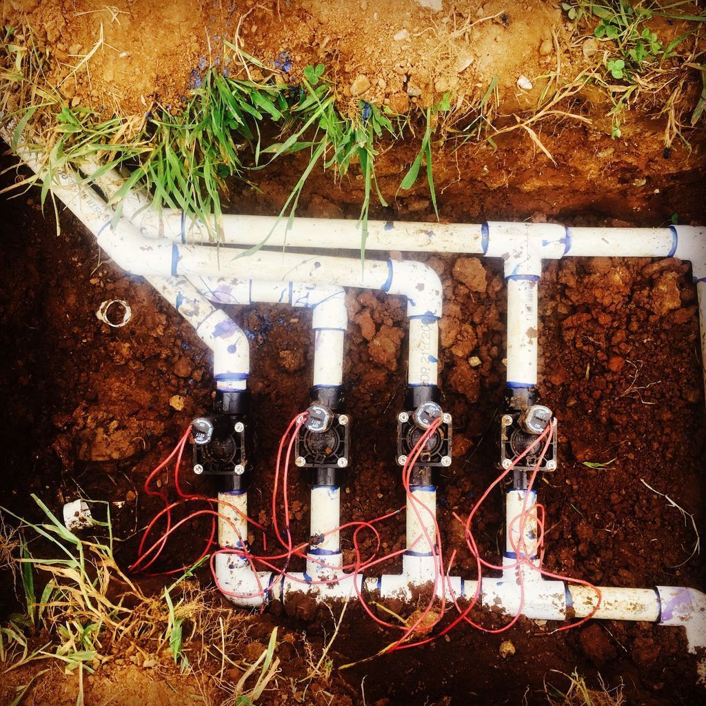 Irrigation valves and PVC pipes installed in a dirt trench with exposed wiring.
