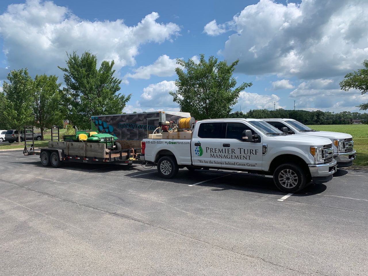 White utility truck with a trailer loaded with supplies parked in a lot under a cloudy blue sky.