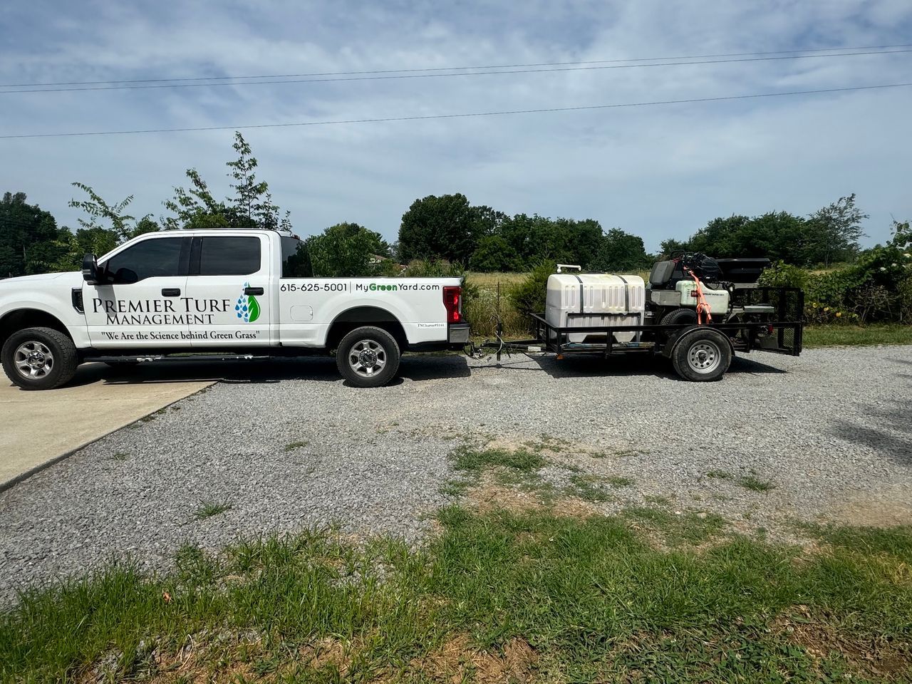 White utility truck towing a tank trailer on a gravel lot under a cloudy sky