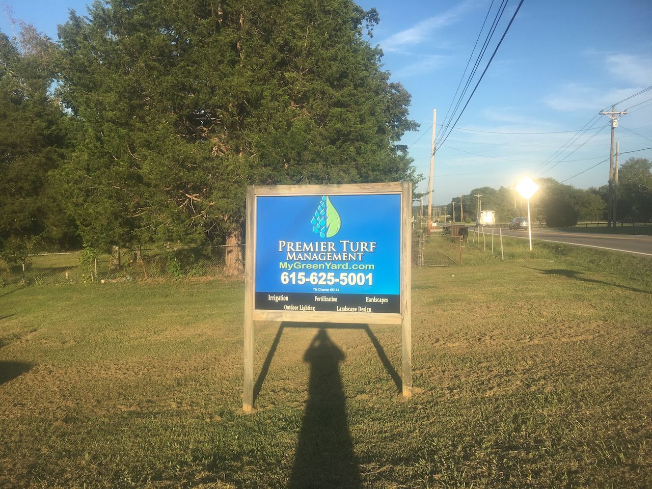 Blue roadside sign in a grassy field with a phone number and a road visible to the right.