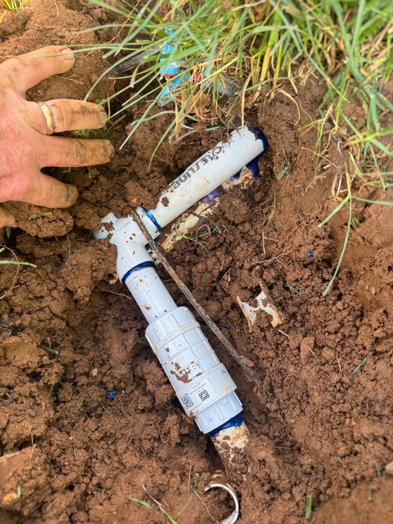 Gloved hand points to broken white PVC pipe in muddy soil near grass roots