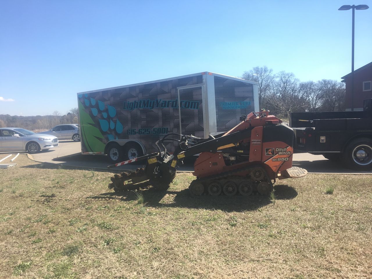 Two small tractors parked on grass beside a branded trailer in a parking lot