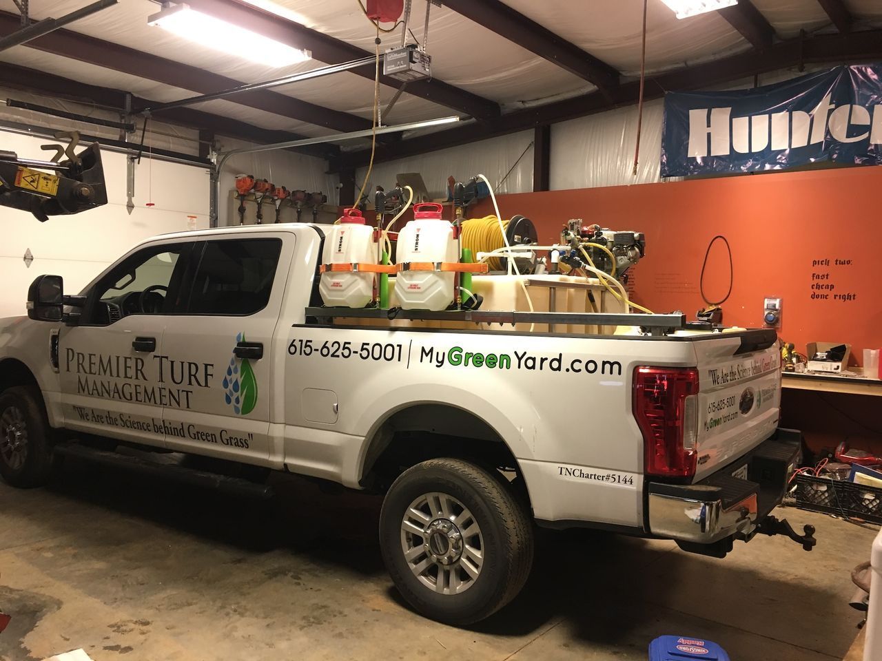 White pickup truck with landscaping equipment parked inside a garage, with buckets and tools in the bed
