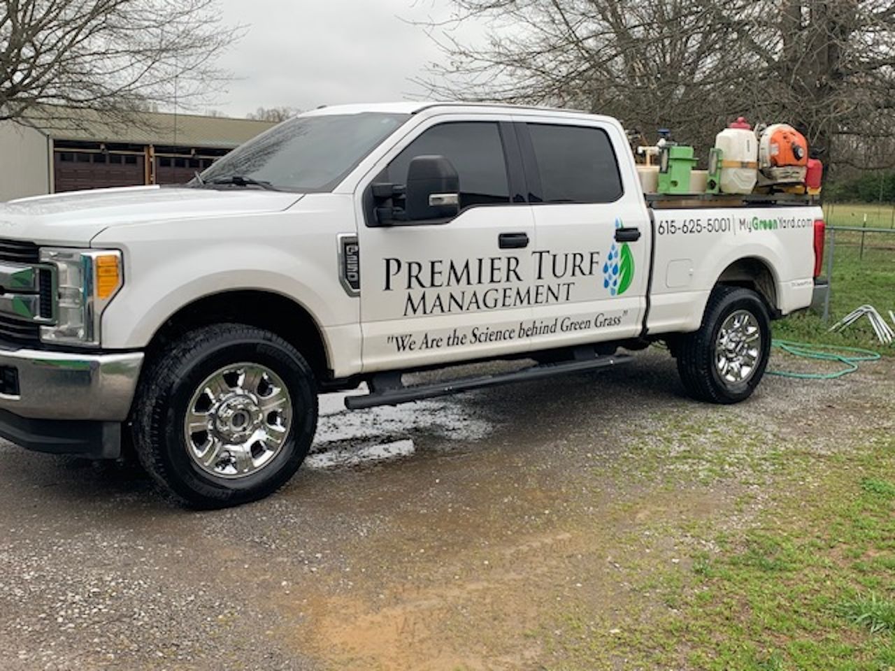 White Premier Turf Management pickup truck parked on gravel, with landscaping equipment in the back.