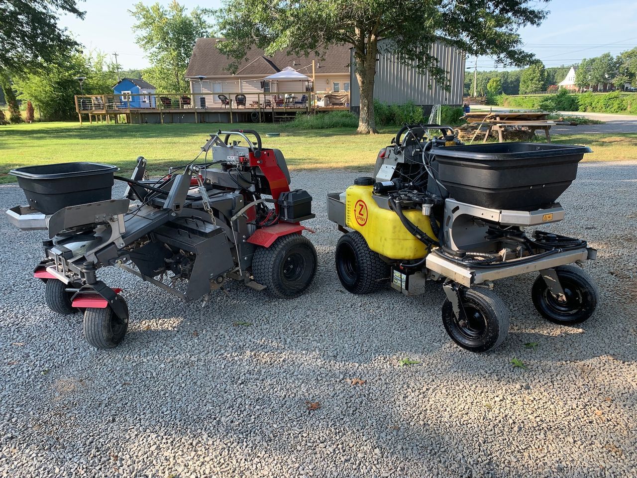 Two riding lawn mowers, one red and one yellow, parked on gravel outdoors near a house.