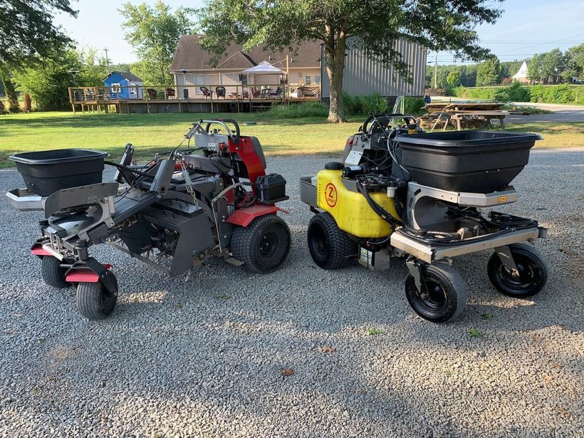 Two riding lawn mowers, one red and one yellow, parked on a gravel driveway in a yard.