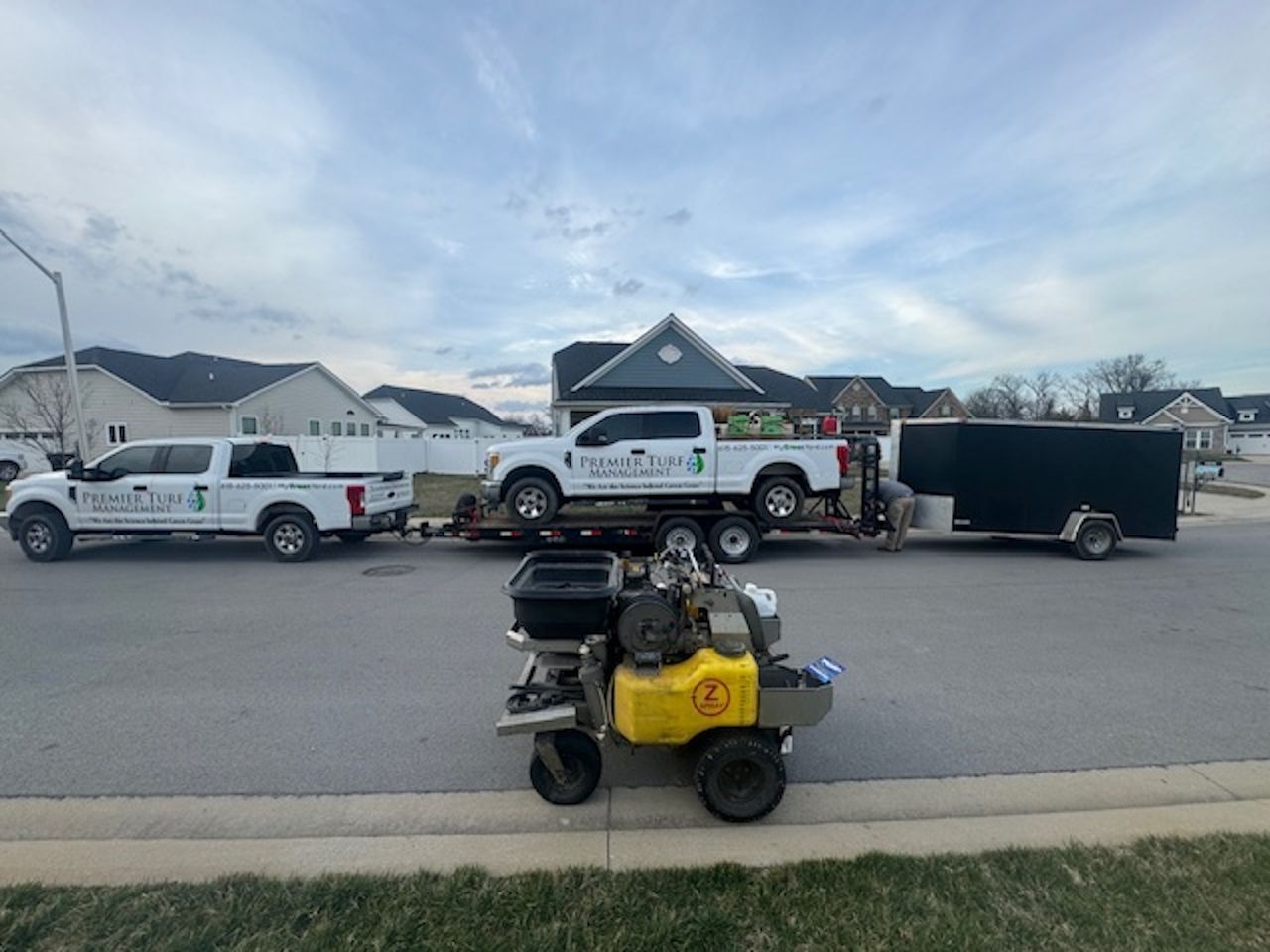 Small yellow mini bike parked in a residential street with work trucks and a trailer in the background