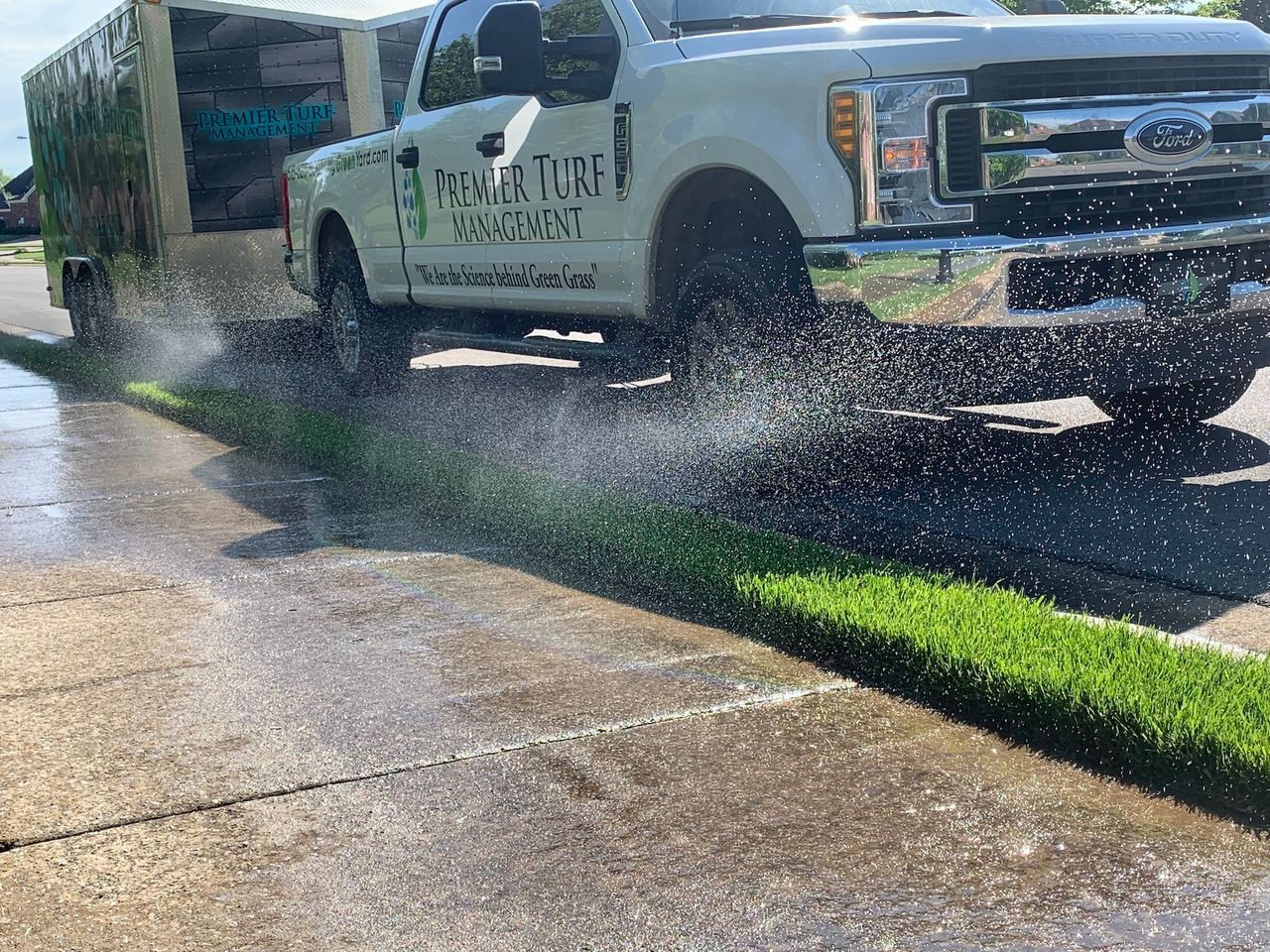 White pickup truck towing a trailer sprays water while driving past a wet curbside lawn
