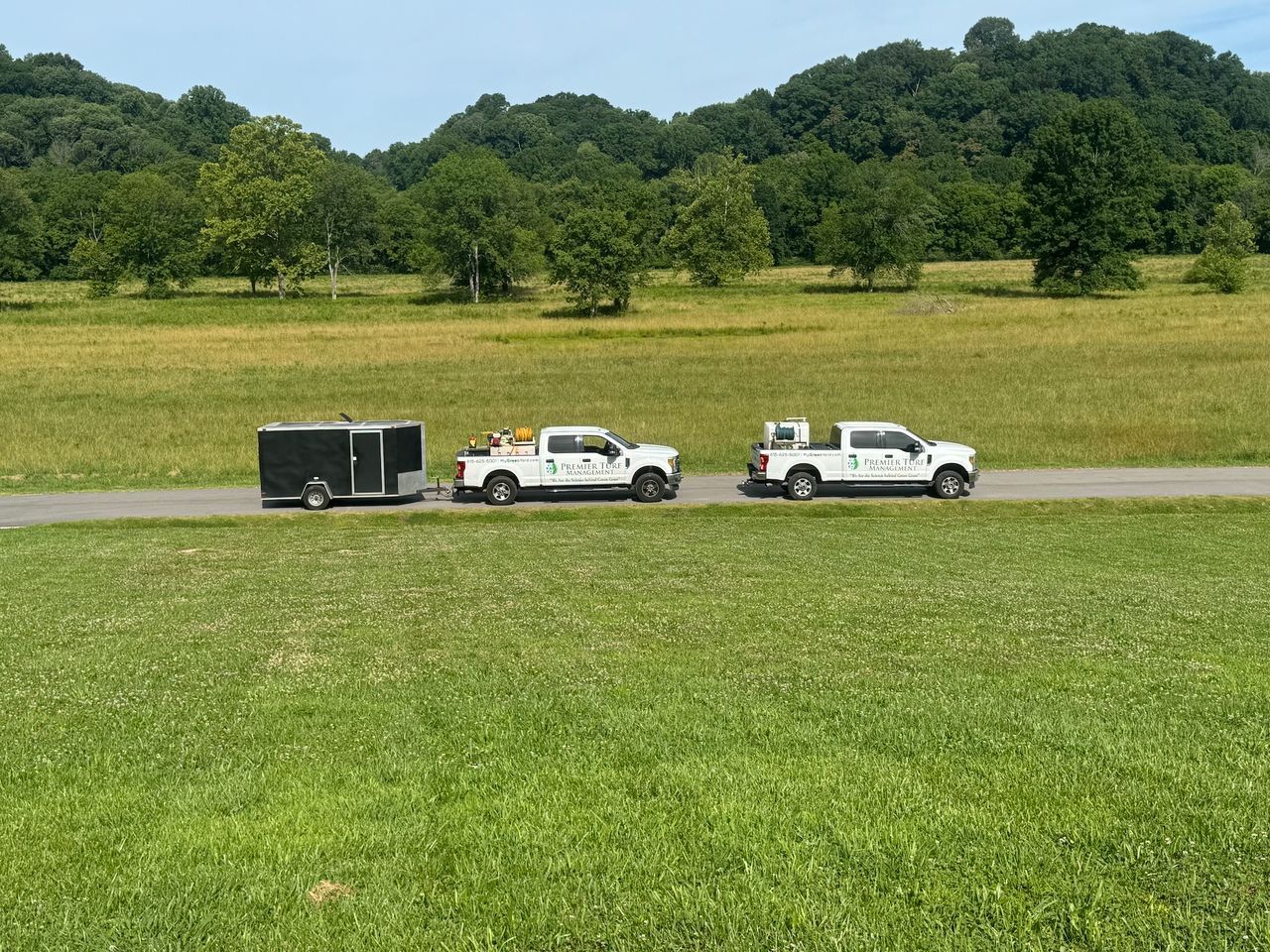 Two white pickup trucks towing a black trailer across a grassy field with trees in the background