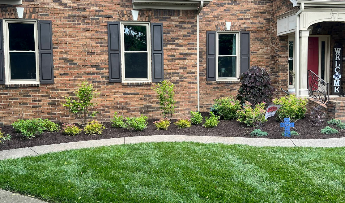 Brick house front garden with black shutters, flower beds, and a curved stone border along the lawn